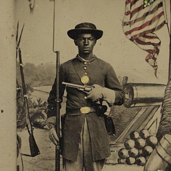 A black soldier standing with musket at his side holding a pistol across his the front of his body.