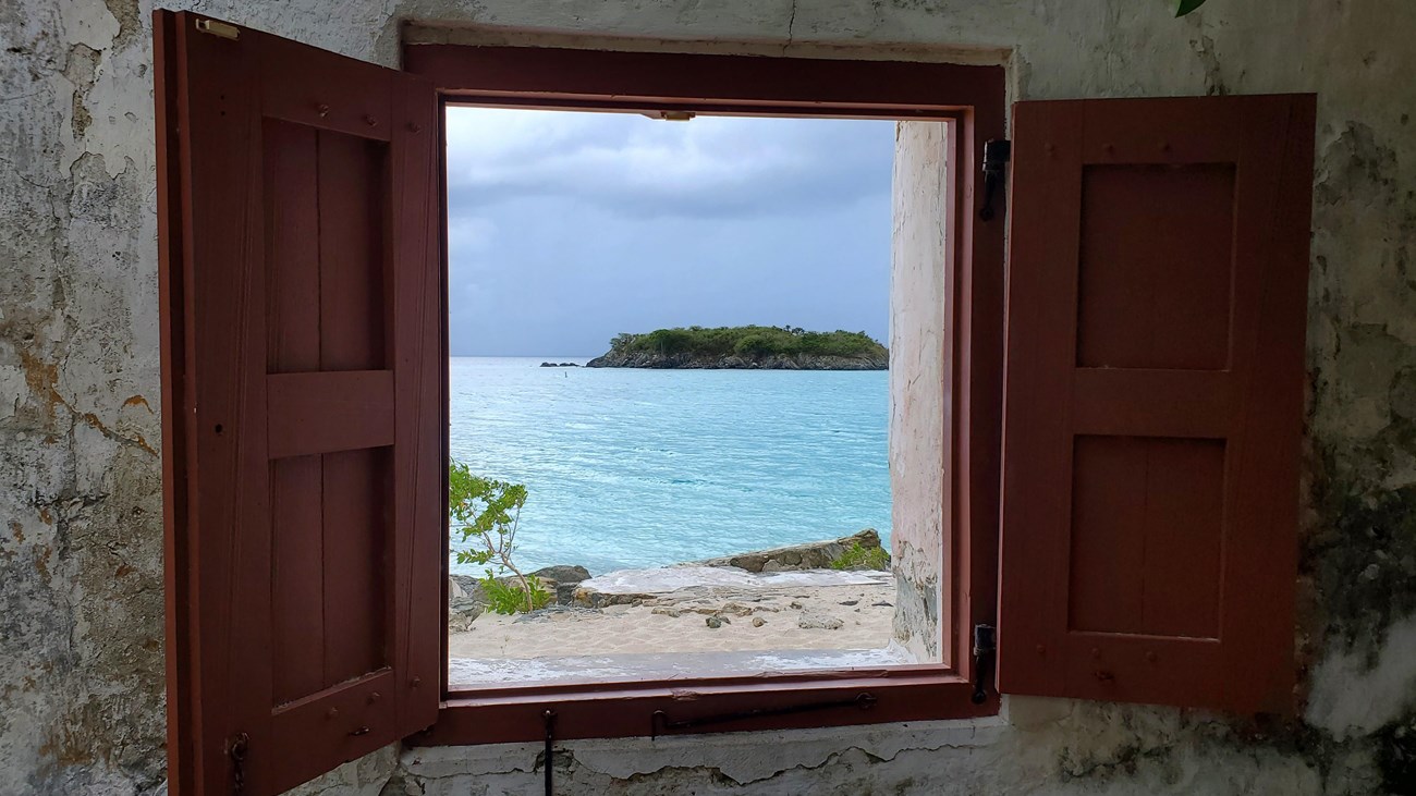 An open window with dark red shutters and to view of a white sand beach and turquoise waters.