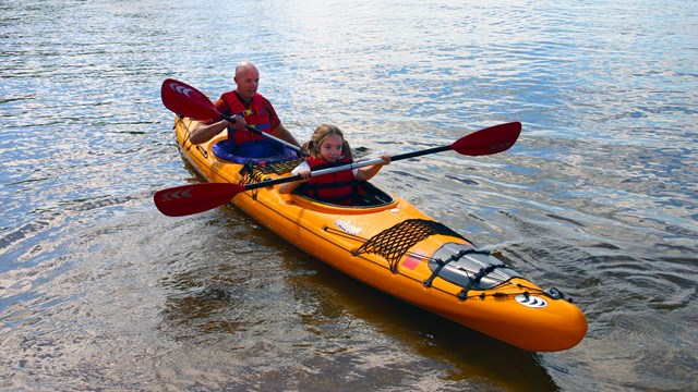 A bright yellow kayak floats near the shore, paddled by a young girl and her father.