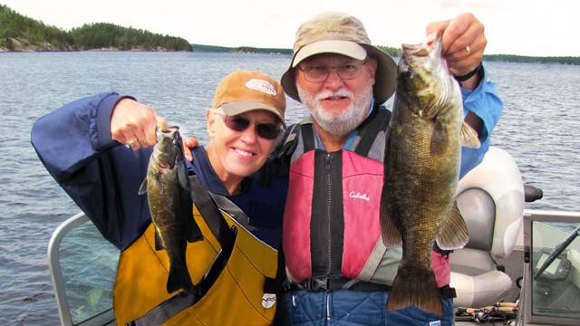 A man and a woman in life jackets stand in a boat on a scenic lake and hold recently caught fish.