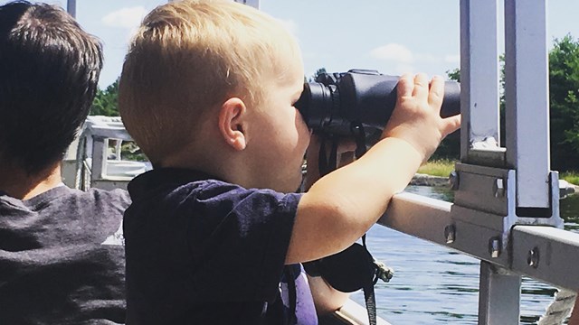 A young child sits in a tour boat and holds a pair of binoculars up to his eyes.