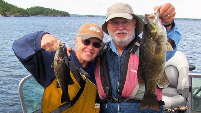 A man and woman stand in a boat upon a scenic lake rimmed by trees. Both hold up Smallmouth Bass.