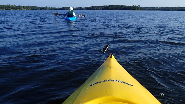 The nose of a bright yellow kayak points outwards towards a kayaker in the distance on a scenic lake