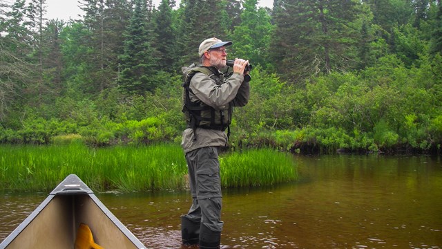 Photograph of man standing in water with canoe nearby holding binoculars.