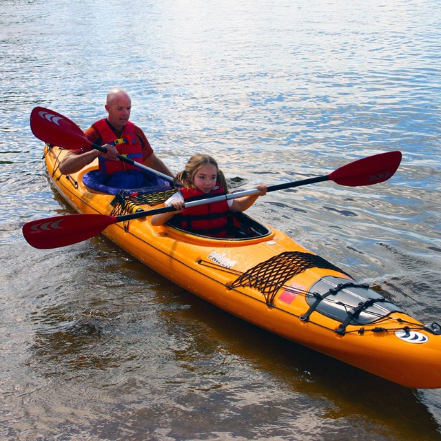 A bright yellow kayak floats near the shore, paddled by a young girl and her father.