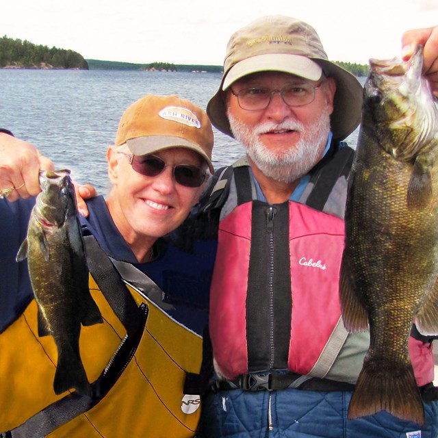 A man and a woman in life jackets stand in a boat on a scenic lake and hold recently caught fish.