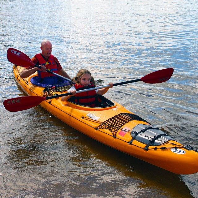 A bright yellow kayak floats near the shore, paddled by a young girl and her father.