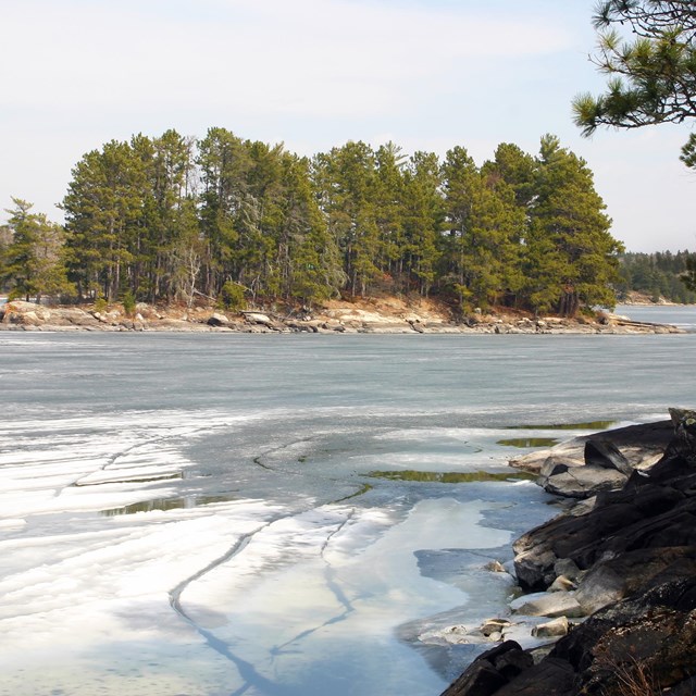 Slushy, gray ice covers the surface of a scenic lake, bordered by rocks and trees on the far shore.