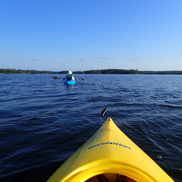 The nose of a bright yellow kayak points outwards towards a kayaker in the distance on a scenic lake