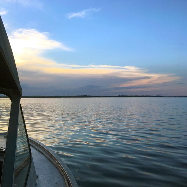 The sun sets behind clouds on the horizon of a calm, scenic lake, seen from the cockpit of a boat.