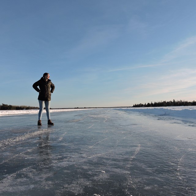 A visitor stands on a wide stretch of cleared ice on the surface of a scenic lake.