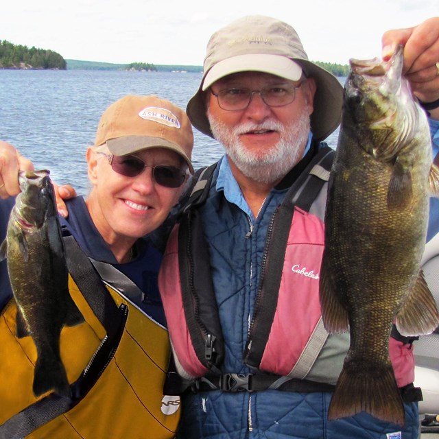 A man and a woman stand in a boat on a scenic lake and hold up two Smallmouth Bass they just caught.