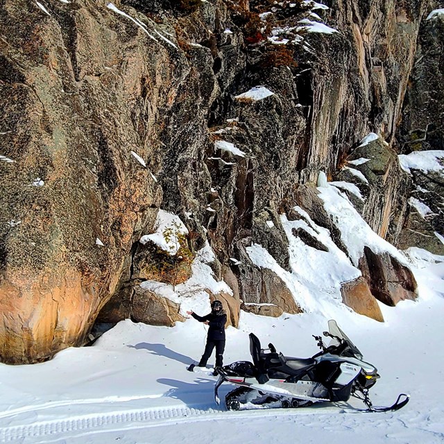 A woman in a snowmobile suit and helmet stands beside a snowmobile and gestures to cliffs behind her