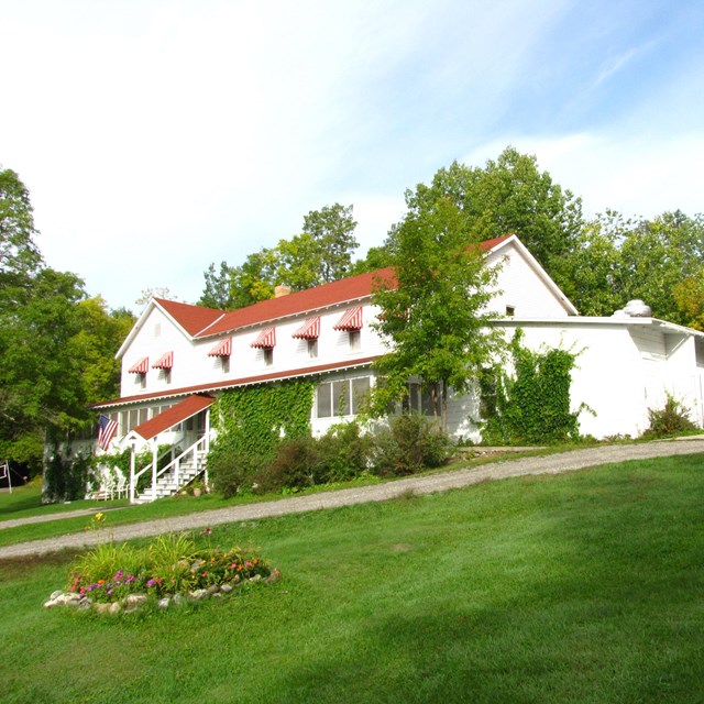 A large, white hotel building with a red roof is surrounded by green grass and thick trees.