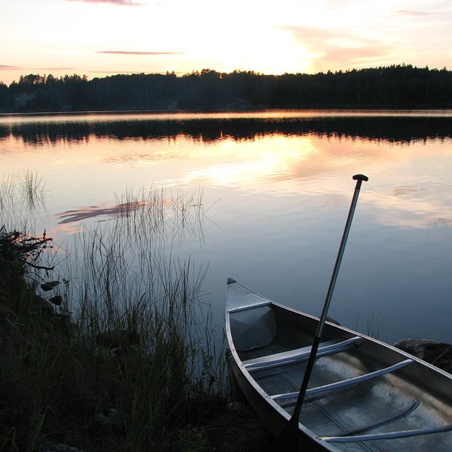 An empty canoe sits on the shore of a small, scenic lake at sunset.