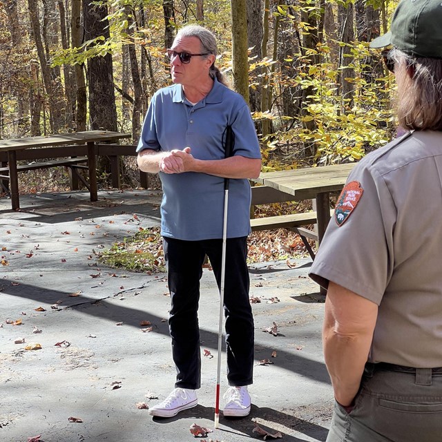 A man wearing sunglasses and holding a white-tipped cane stands with his hands clasped.