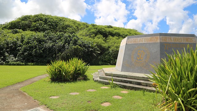 A six-sided concrete monument in front of a mountain covered in vegetation.