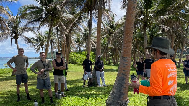 NPS staff speaking to volunteers at Asan Beach Park, under coconut trees by the ocean