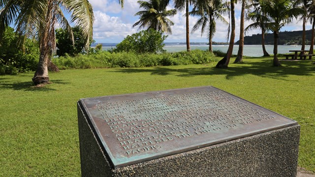 Bronze plaque set on a concrete cube. In the background are palm trees and the ocean.