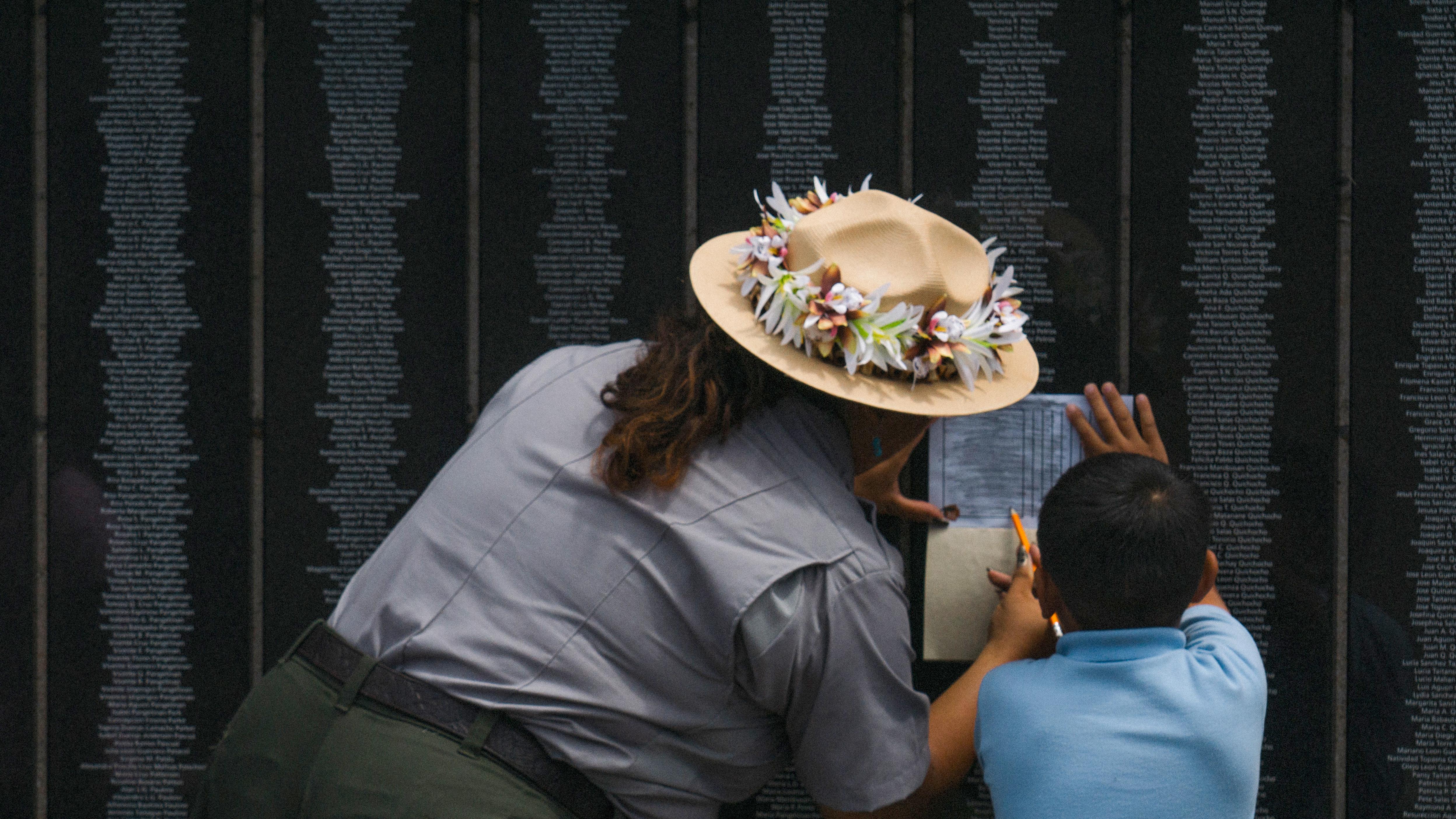 Woman with a flower wreath around a ranger hat and a young boy stand at a wall of names.