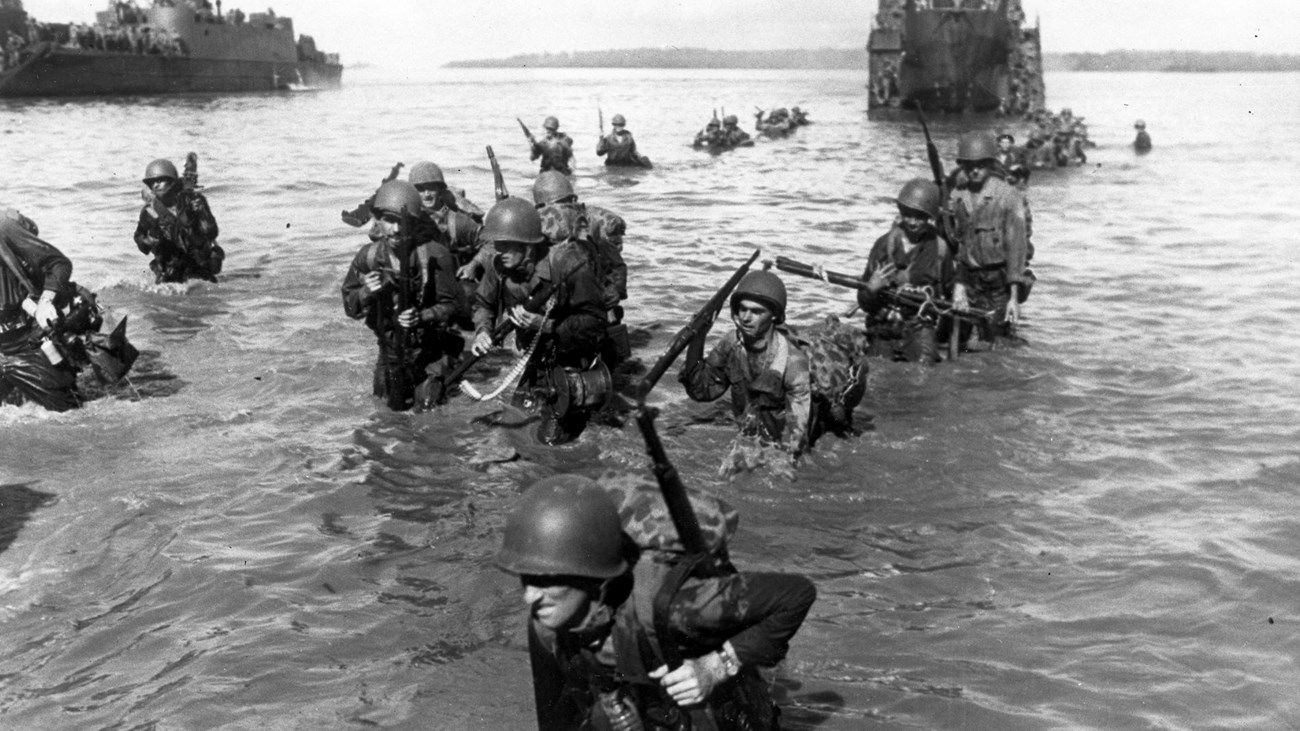Uniformed soldiers carrying their packs and weapons wade through the water from an amtrac.