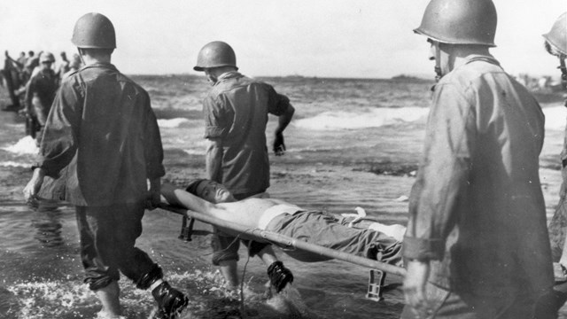 Four soldiers carry an injured man on a stretcher through the water
