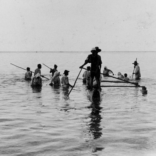 Two men standing on an outrigger canoe. 10 men stand in chest deep water around it.