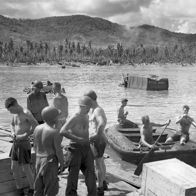 Group of topless soldiers loading supplies from a boat to an island.