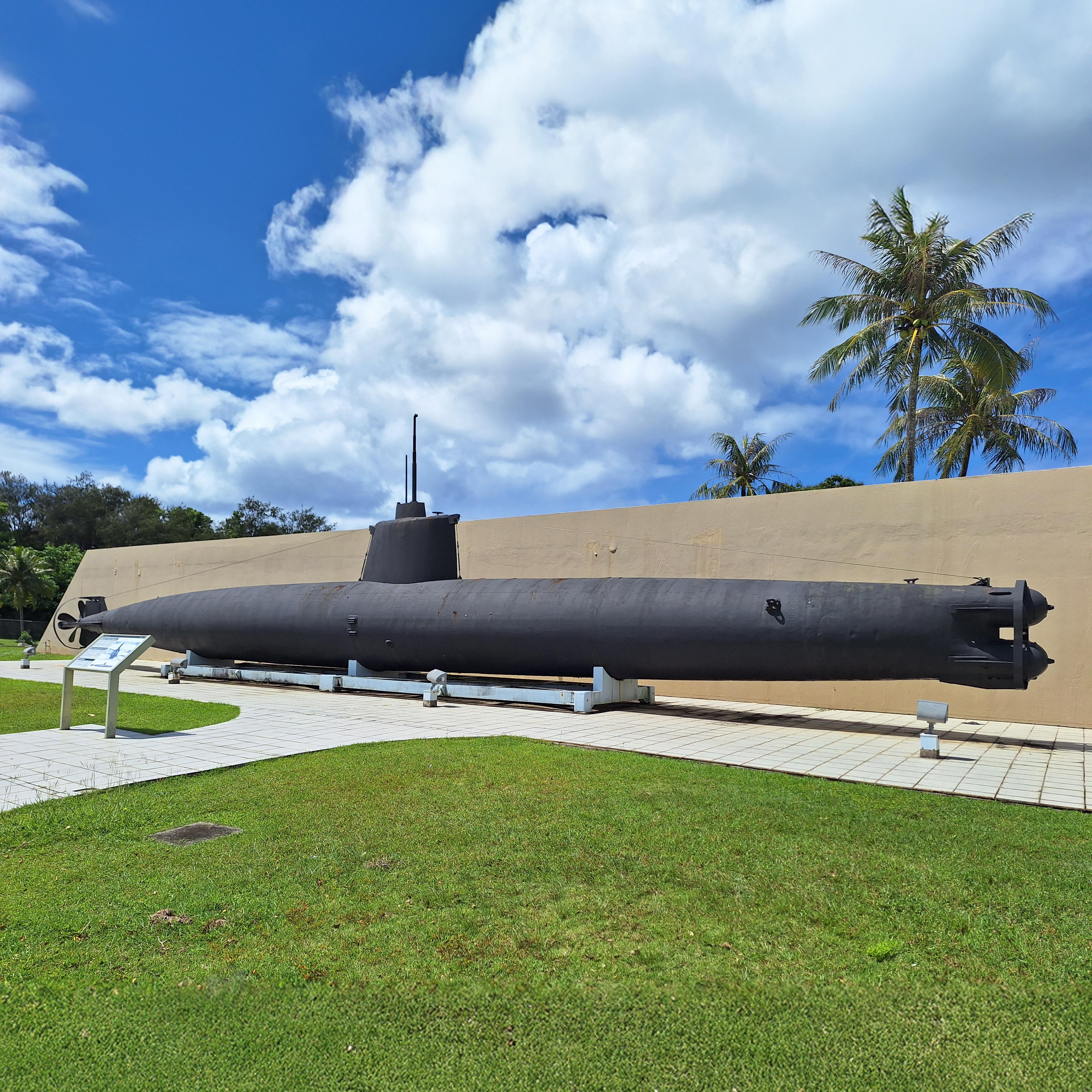 A large black submarine with a periscope resting on a concrete plinth in front of a concrete wall.