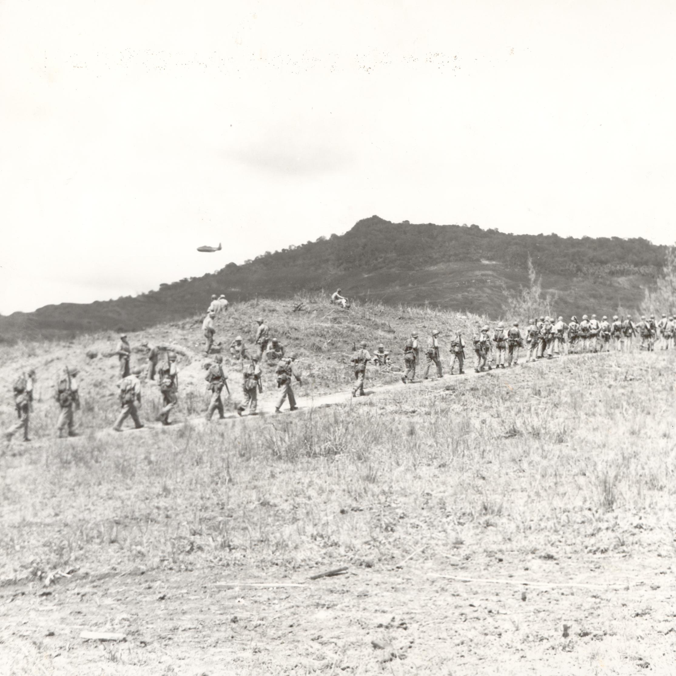 Soldiers march in a single file across a barren landscape, with rugged hills in the background.