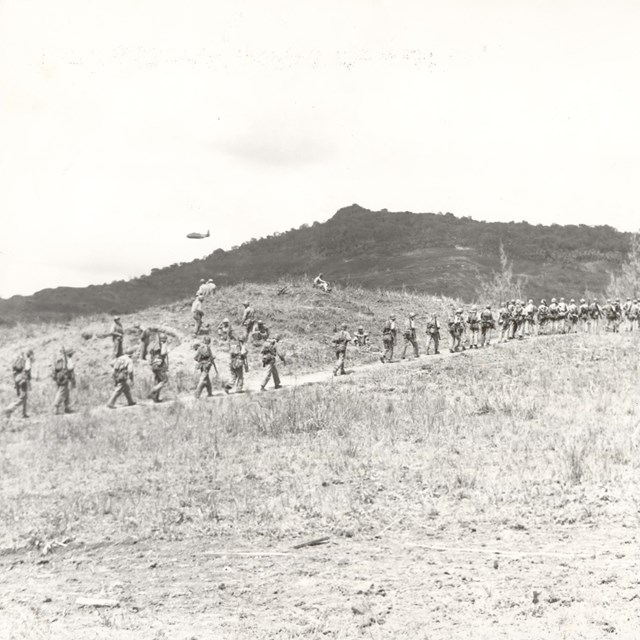 Soldiers march in a single file across a barren landscape, with rugged hills in the background.