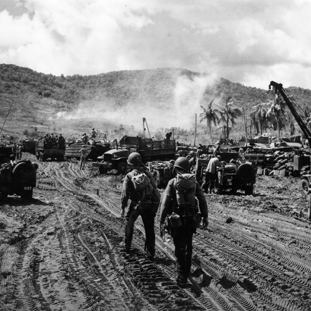 Soldiers in helmets and gear navigate a muddy area with military vehicles and equipment.