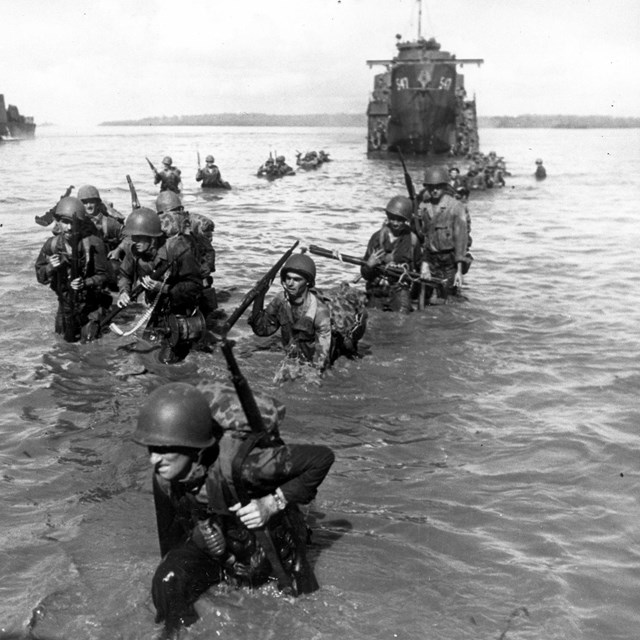 Uniformed soldiers carrying their packs and weapons wade through the water from an amtrac.