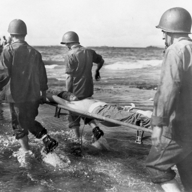 Four soldiers carry an injured man on a stretcher through the water