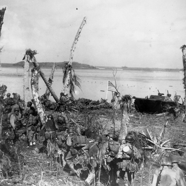 Line of soldiers marching on a bombed out beach next to an American flag
