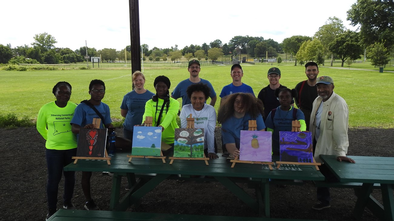 A group of people standing in a park with several paintings on easels in front of them.