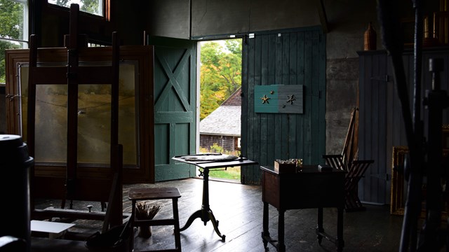 A small dark room with the door open to an outside space. The room is filled with tables and easels.