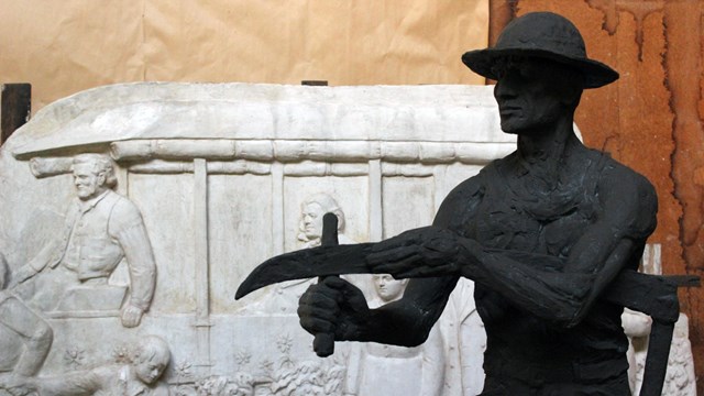 A silhouette of a statue holding a scythe in front of a large white plaster cast of a wagon. 