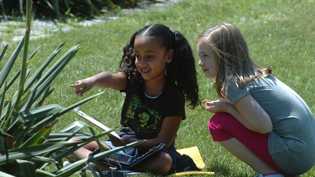 Two young visitors sit on a grassy lawn and point at a large green plant.