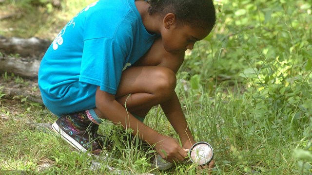 A young girl in a blue shirt is crouched down and looking at the ground with a magnifying glass