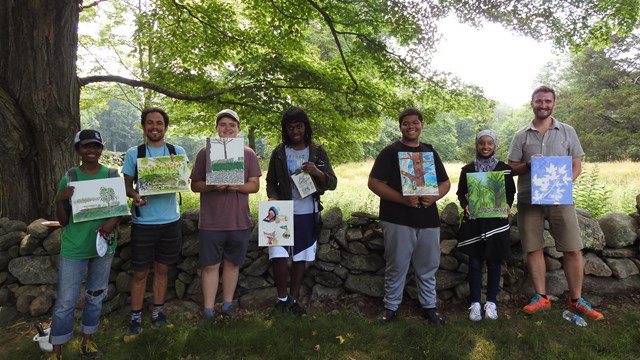 a group of people smile at the camera outside in a shady field holding artwork