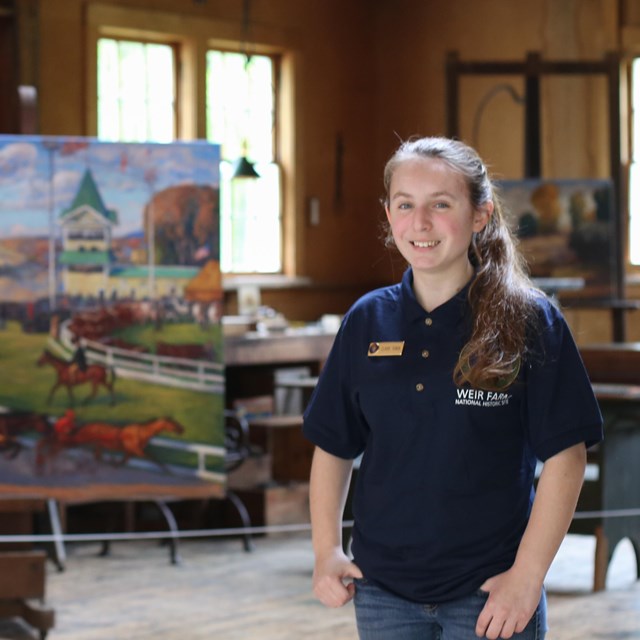 A young person in a blue volunteer shirt stands in front of a painting.