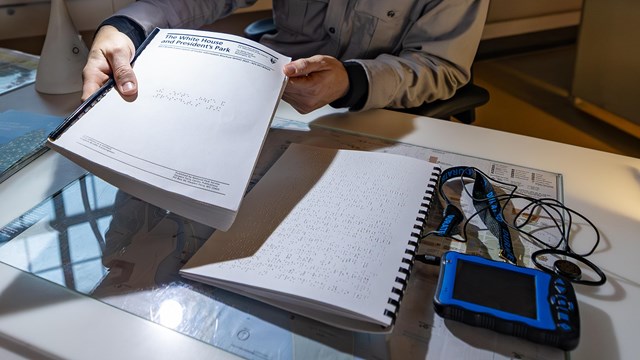 A park ranger holds a braille booklet next to an audio description device.