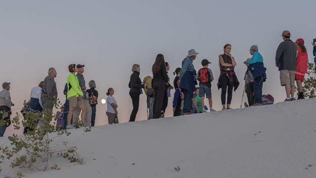 a large crowd of people stand at the top of a white sand dune. The full moon can be seen in a gap.