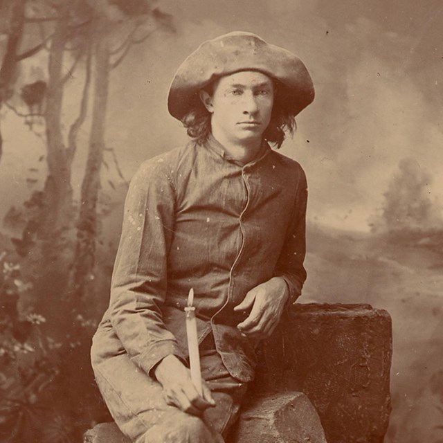 black and white photo of a young man with a wide brimmed hat holding a candle sitting on a rock