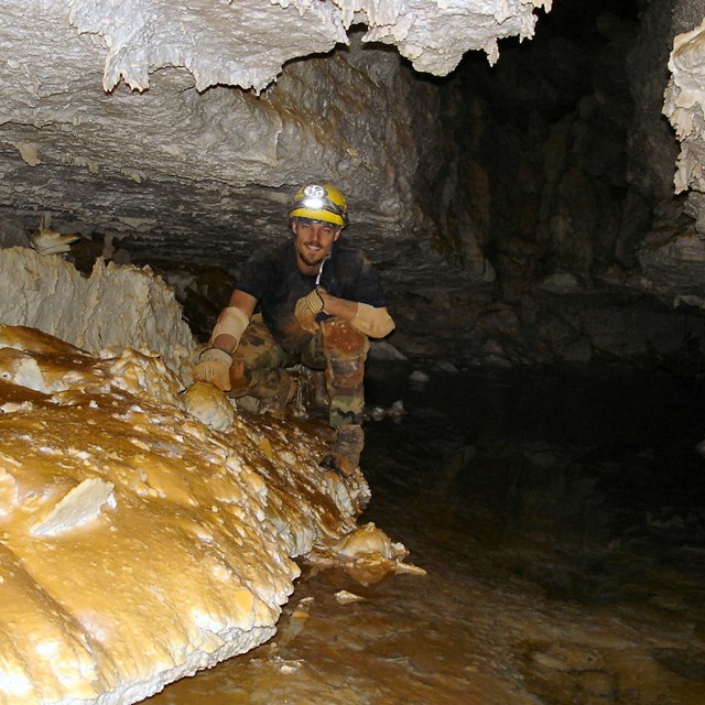 a man in caving gear crouching in a low cave passage next to a shallow clear pool of water