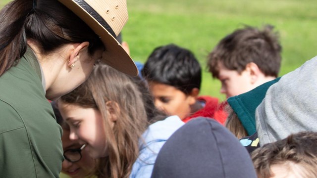 The back of a park ranger as she leans down to help a group of elementary school children. 