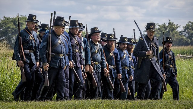 Men dressed in Civil War Uniforms stand in a straight line holding muskets at their sides. 