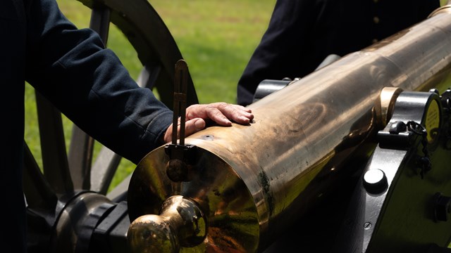 A man out of frame dressed puts his hand on the back of an artillery piece. 