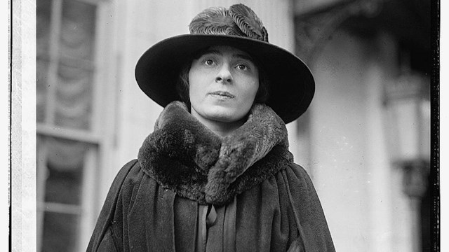 A white woman dressed in black cape, fur collar, and hat is looking up slightly